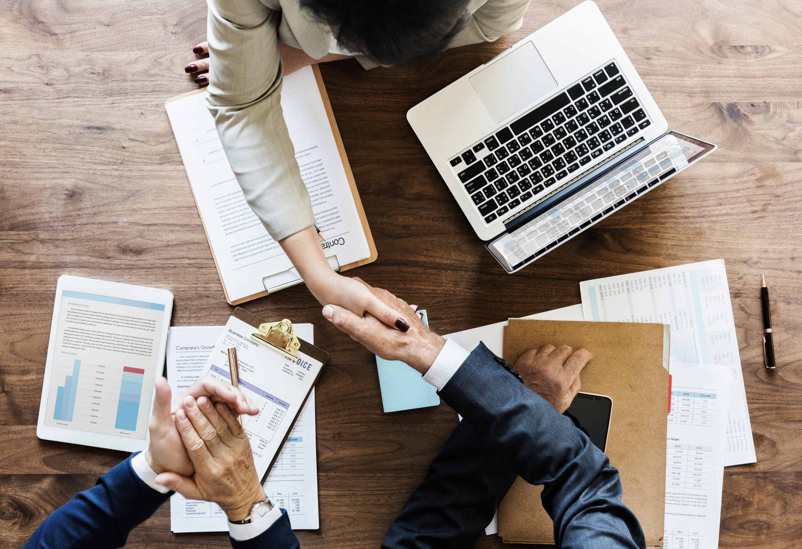 Duas pessoas apertando as mãos em uma mesa de trabalho com documentos, gráficos, laptop e tablet, simbolizando acordo ou negociação.